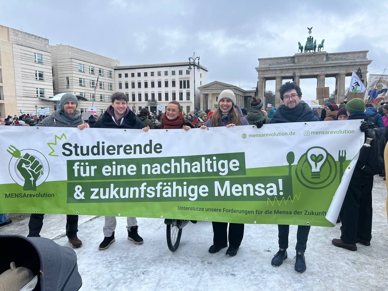 Ein Foto mit junge Menschen, die vor dem Brandenburger Tor in Berlin mit dem Banner der Mensarevolution stehen.